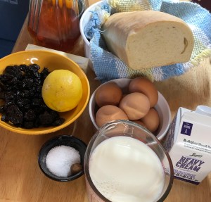 Photo of ingredients. Loaf of bread on a blue, yellow, and white checked cloth, bowl with eggs, bowl with dried cherries and a lemon, bowl with salt and half a nutmeg. a container of heavy cream, and a measuring cup with milk.