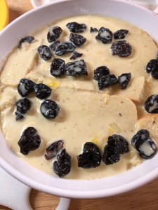 Pudding mid-assembly. White baking dish with a layer of sliced bread with dried cherries sprinkled on the top. Custard mixture poured over the layers.
