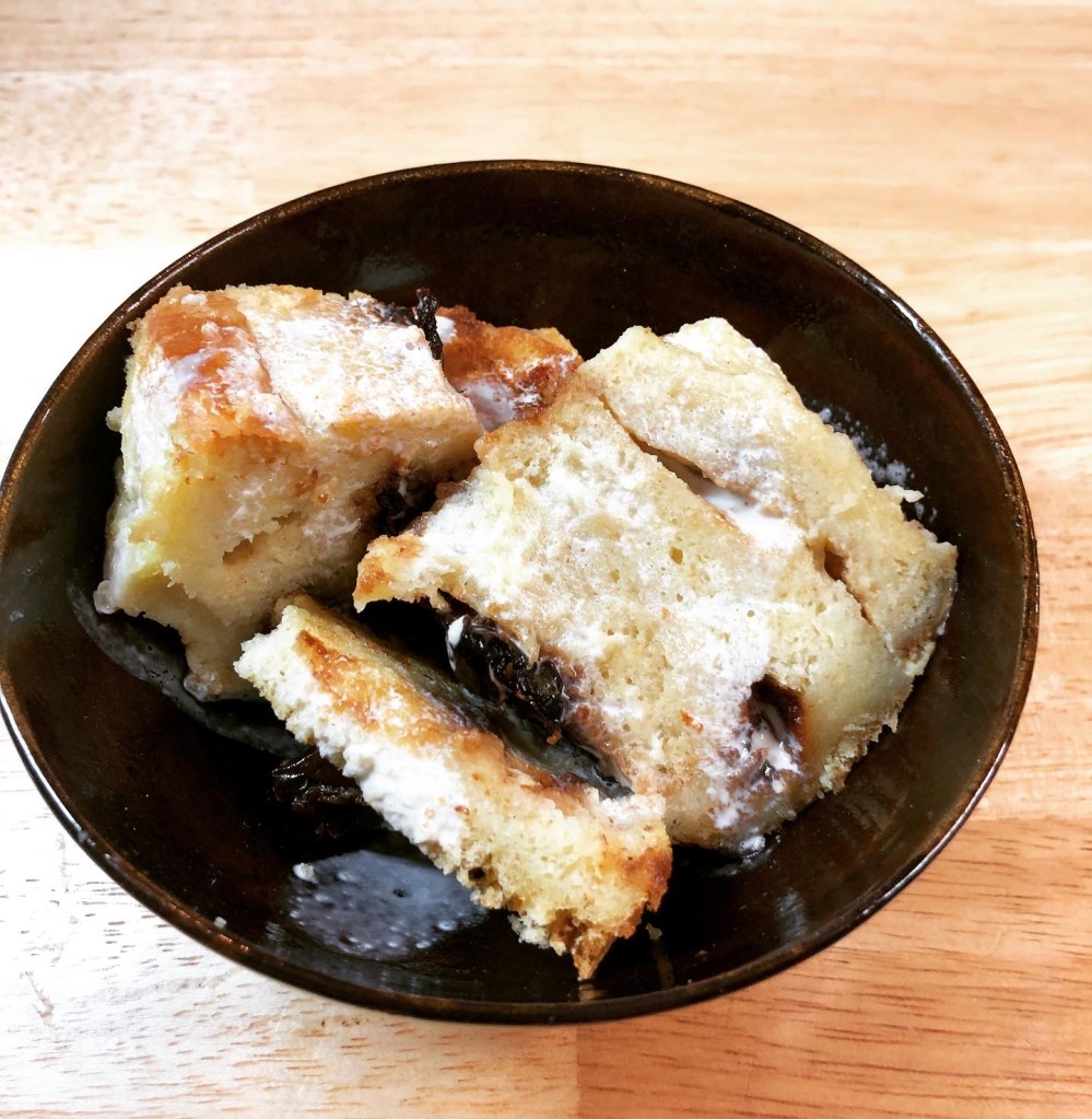 Brown pottery bowl with a serving of the pudding. Layers of bread held together with cooked custard with dried cherries. Drizzled with cream.