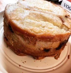 Pudding turned out from baking dish onto plate. Large cylinder of layered bread held together with set custard. Glimpses of dried cherries are visible in the layers.